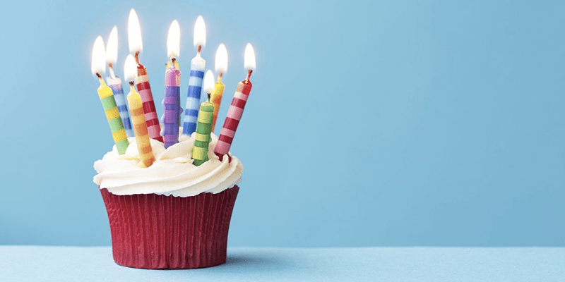 A red velvet cupcake with creme cheese icing has ten lit candles in it. The candles are green, red, orange, and purple. The cupcake is setting on a light blue table with a light blue wall in the background.